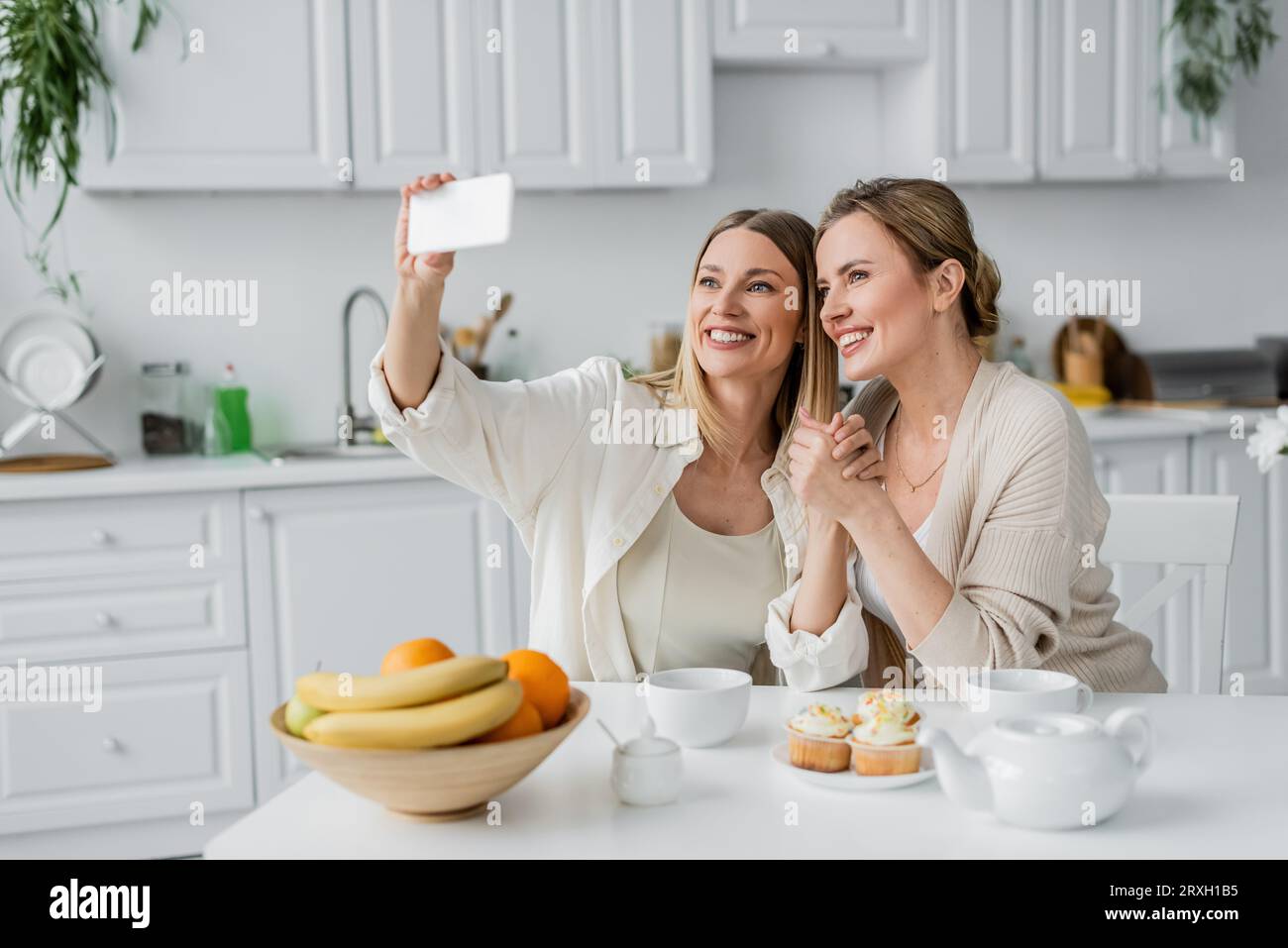 Klassische Schwestern in trendigen Pastell-Cardigans machen Selfie am Tisch in der Küche, Familienbindung Stockfoto