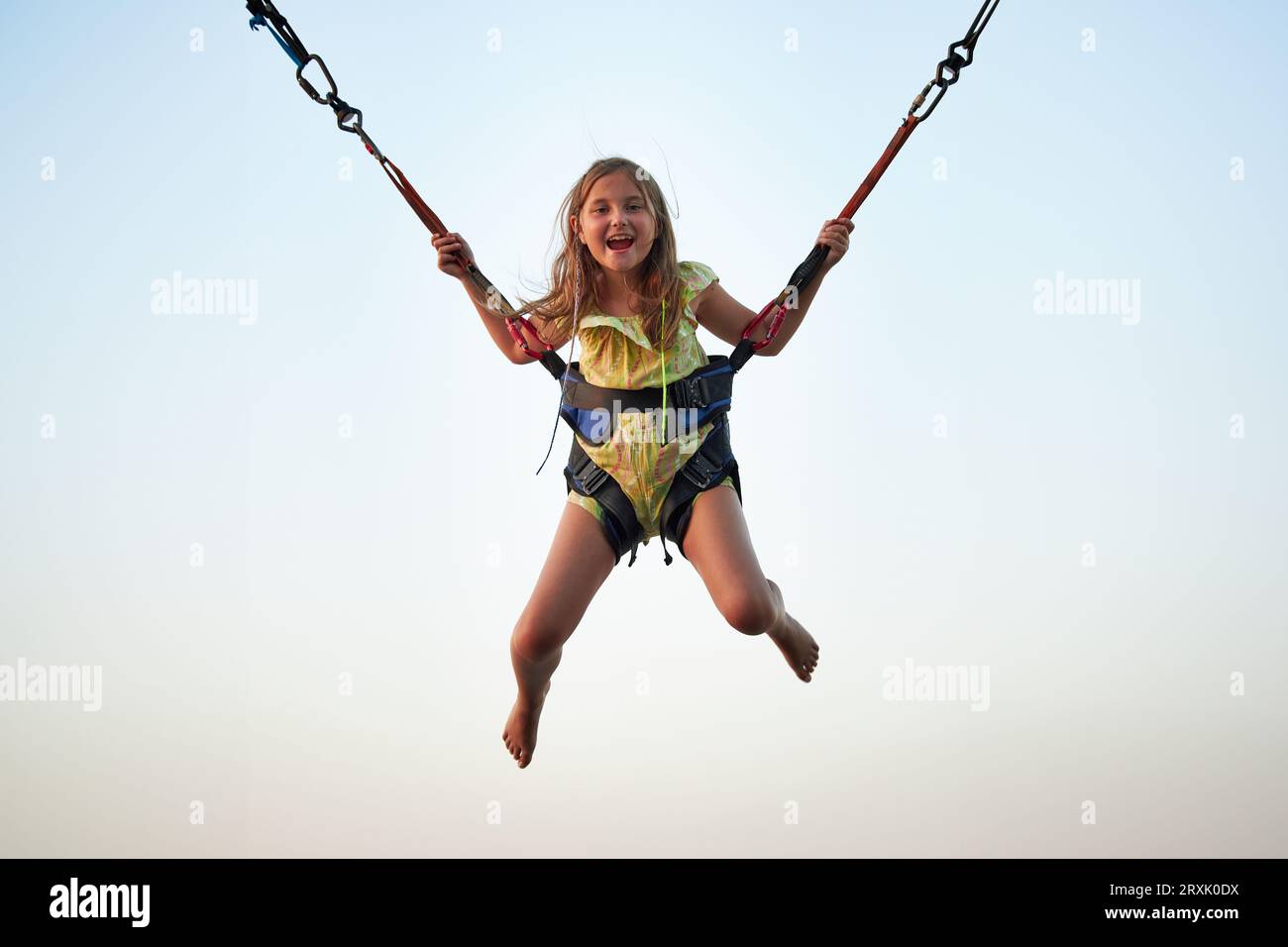 Bungee-Jumping auf Trampolin. Kleines Mädchen hüpft auf Bungee-Jumping im Vergnügungspark auf Sommerferien. Kind fliegt über den blauen Himmel, während es weiterspringt Stockfoto