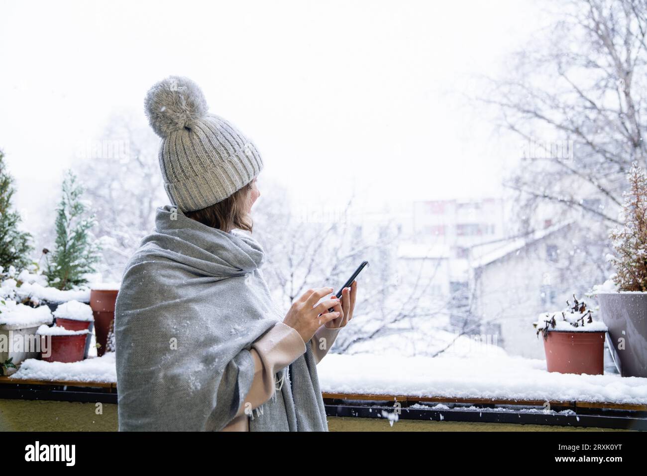 Eine Frau in einem warmen Hut und einer Decke hält das Smartphone in den Händen. Winterlandschaft mit Schneefall im Hintergrund. Stockfoto