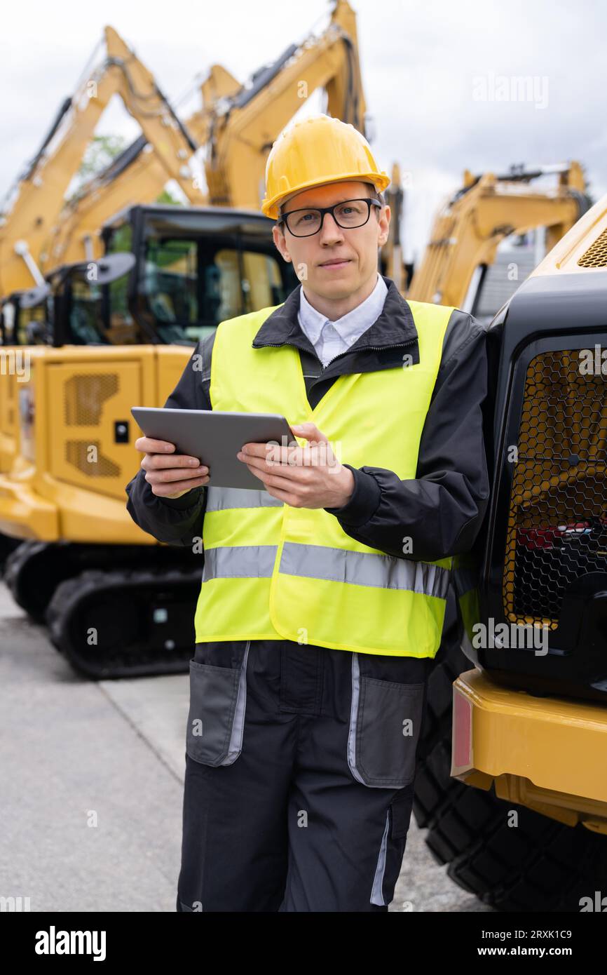 Ingenieur mit Helm und digitalem Tablet steht neben Baubaggern. Stockfoto