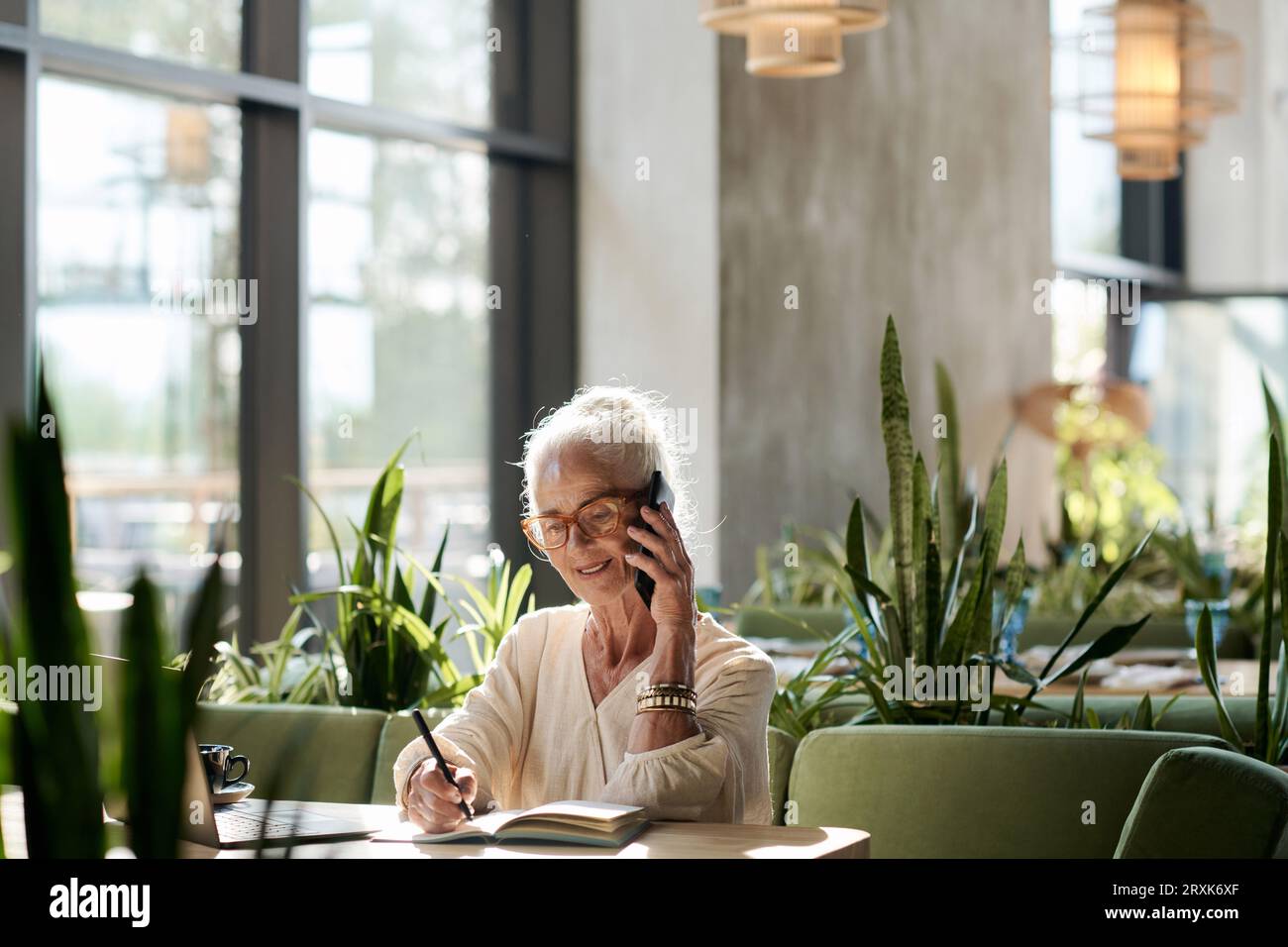 Ältere Frau, die sich während ihrer Arbeit im Café mit dem Handy unterhalten und Notizen in ihrem Notizblock macht Stockfoto