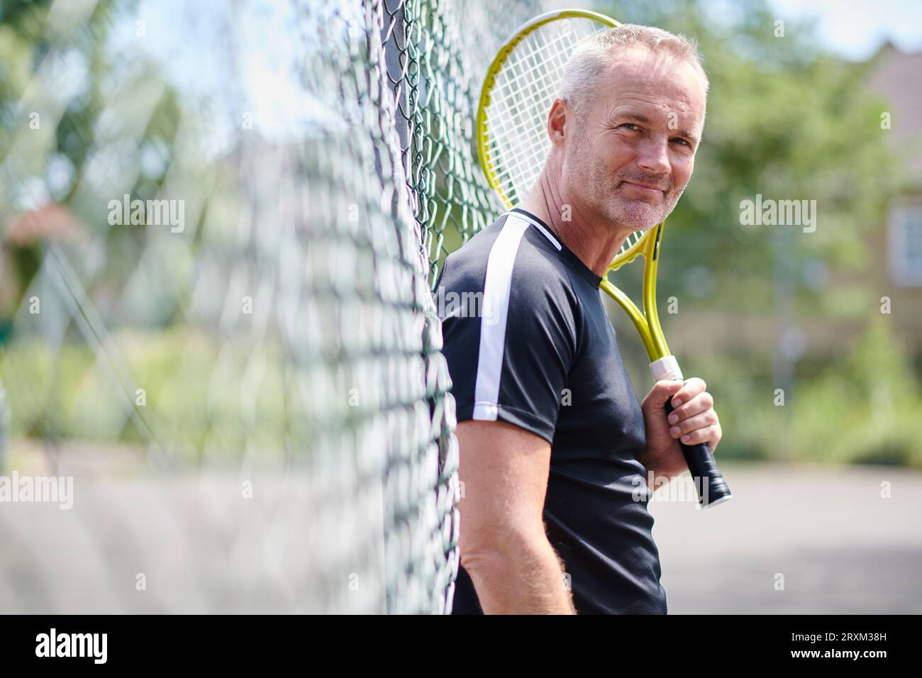 Ein erwachsener Mann, der Tennisschläger hält und sich am Zaun lehnt Stockfoto