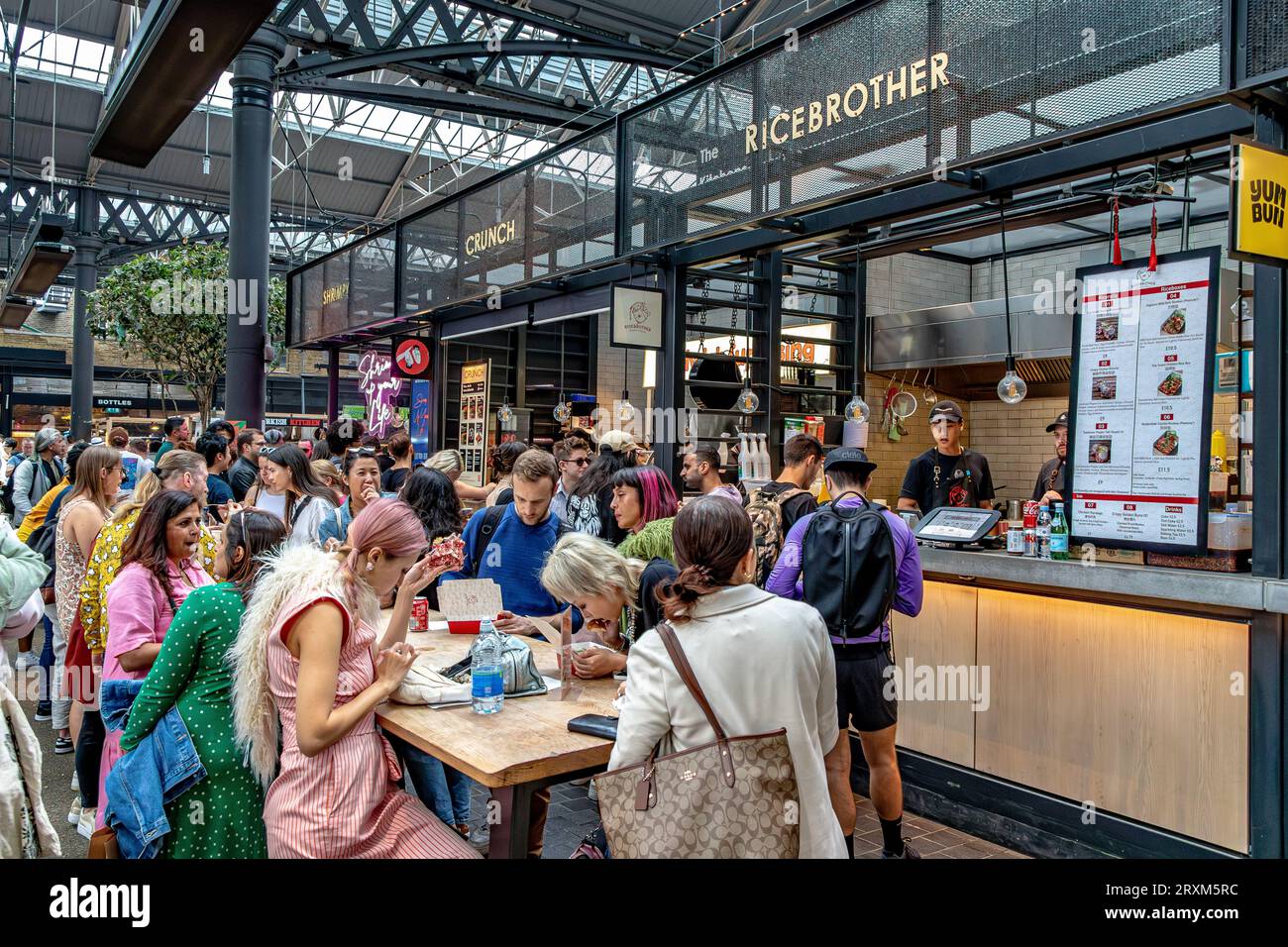 Die Leute, die sich an Tischen in der Nähe verschiedener Restaurants im Old Spitalfields Market, London E1, hinsetzen, essen Essen Stockfoto