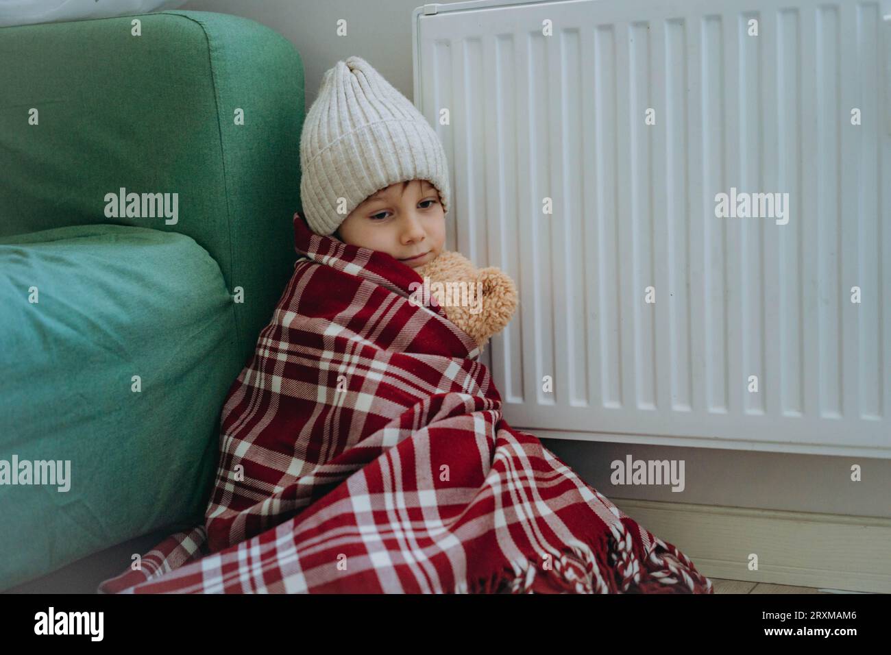 Niedlicher kleiner Junge, der mit einem Karomuster umwickelt ist und Strickmütze trägt, sitzt am Heizer und umarmt den Teddybären. Kälte zu Hause, Energiekrise. Energiearmut Stockfoto