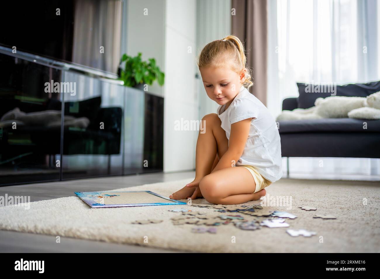 Das kleine blonde Mädchen sitzt zu Hause auf dem Teppich und sammelt Rätsel Stockfoto