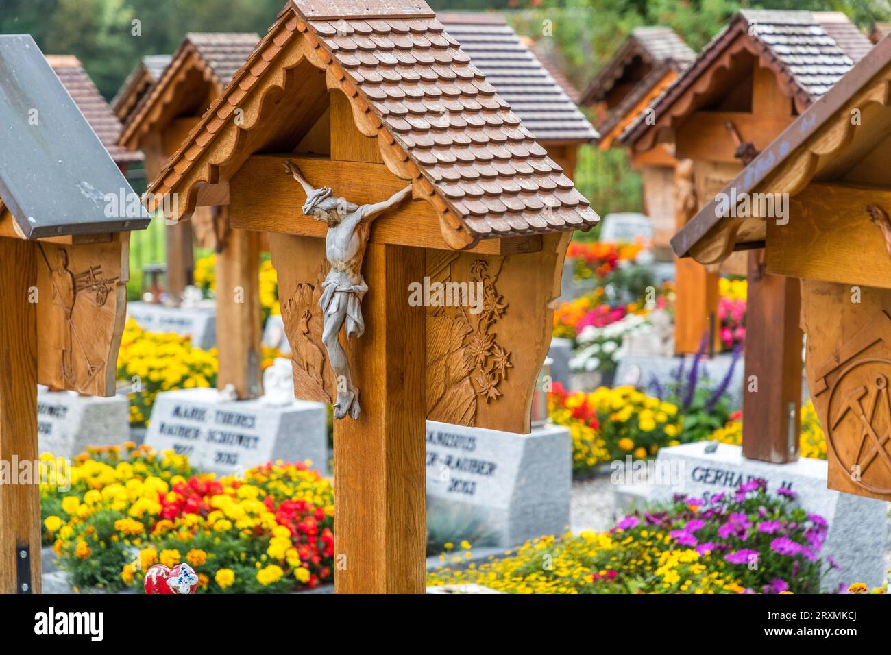 Eine Reihe versetzter Grabkreuze steht auf dem Friedhof hinter der Kirche St. Stephan in Jaun, mit einer leicht bemalten Christusfigur im Vordergrund vor einem Relief, das aus diesem Winkel nicht deutlich sichtbar ist. Jedes der individuell geschnitzten Holzkreuze trägt auf der Rückseite ein Relief, das den Beruf oder das Hobby der darunter begrabenen Person darstellt, eine Tradition, die 1948 vom Autodidakten Walter Cottier begründet und seit 1988 von seinem Cousin Reynold Boschung fortgeführt wurde. Bunte Blumenbeete und Grabsteine säumen die Reihe der Kreuze, Kanton Freiburg, Schweiz. Stockfoto