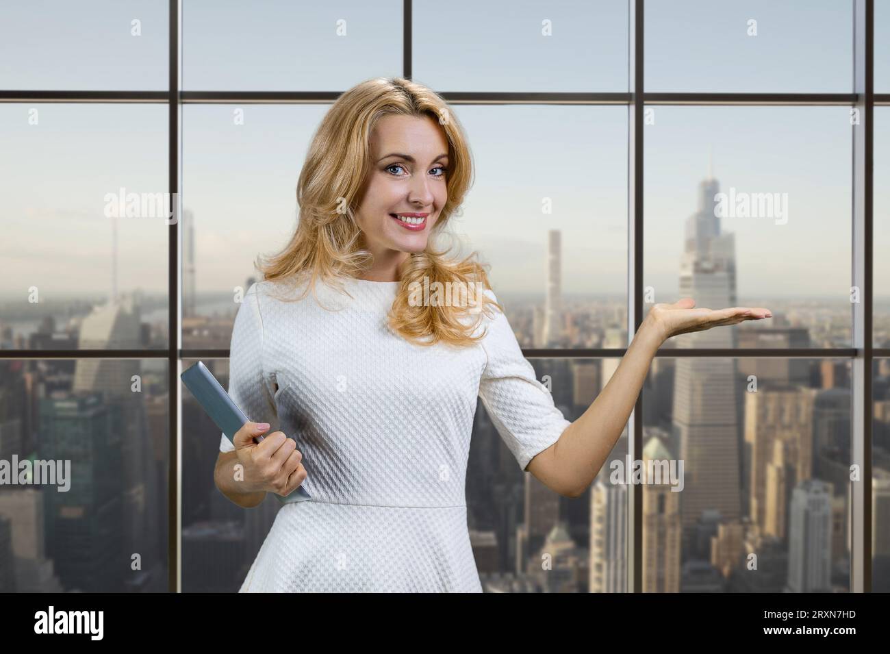 Lächelnde blonde, reife Frau mit Werbefläche für Tablets. Karierte Fenster mit Stadtbild im Hintergrund. Stockfoto