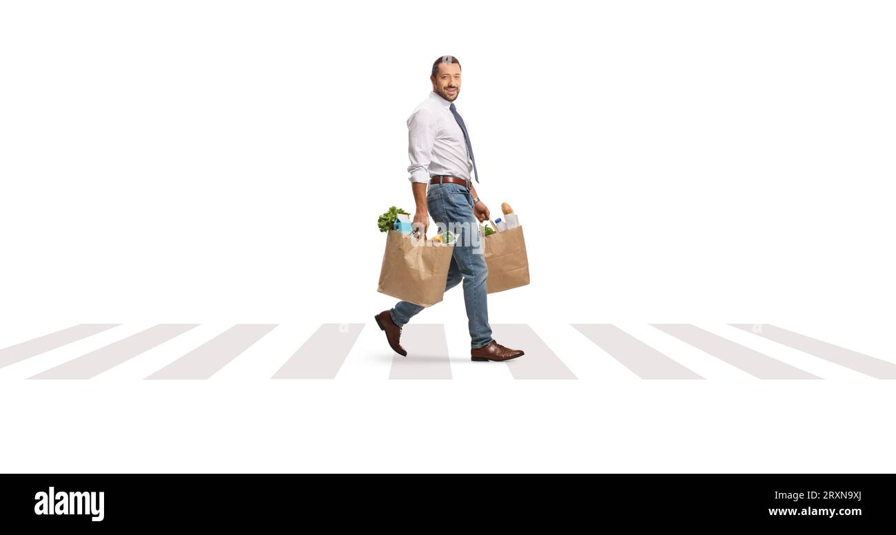 Aufnahme in voller Länge eines Geschäftsmannes, der auf einer Fußgängerüberquerung mit Einkaufstaschen spaziert und die Kamera isoliert auf weißem Hintergrund betrachtet Stockfoto