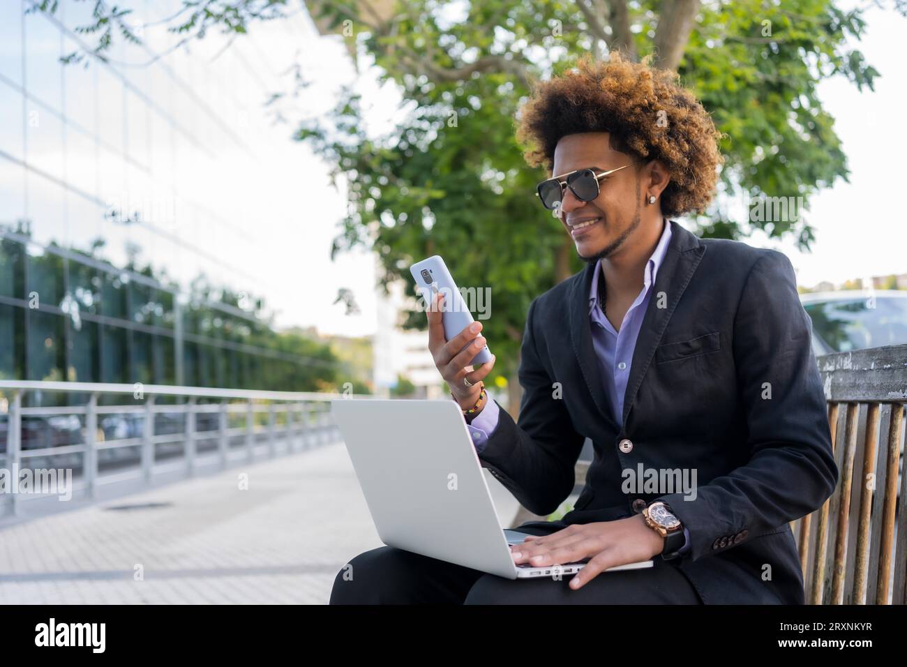 Ein cooler Geschäftsmann, der mit Handy und Laptop arbeitet, während er auf einer Parkbank in der Stadt sitzt Stockfoto