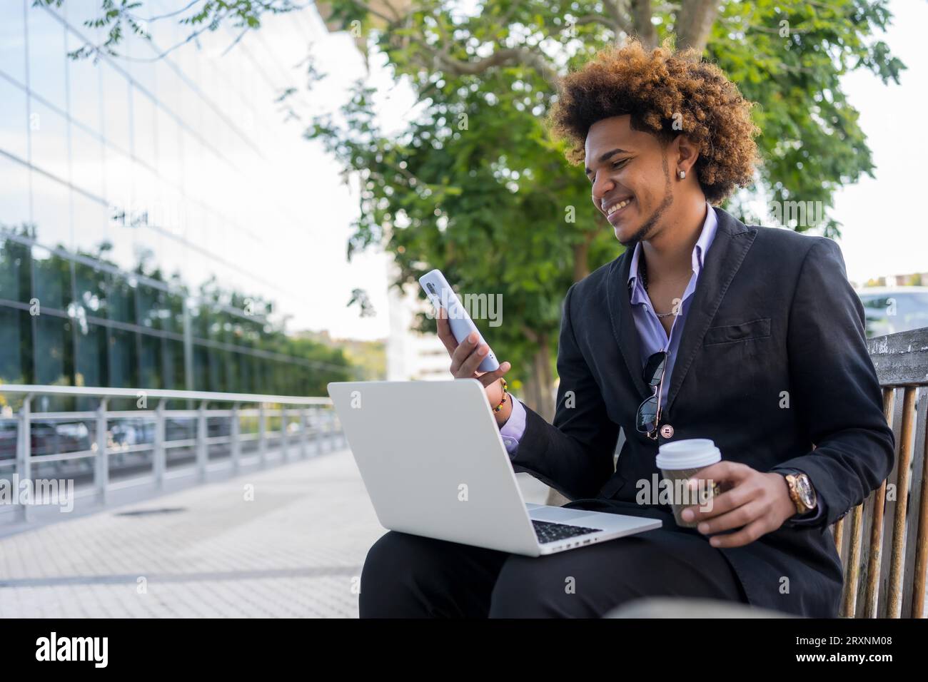 Cooler afroamerikanischer Geschäftsmann, der mit Laptop und Handy arbeitet und morgens auf der Straße sitzt Stockfoto