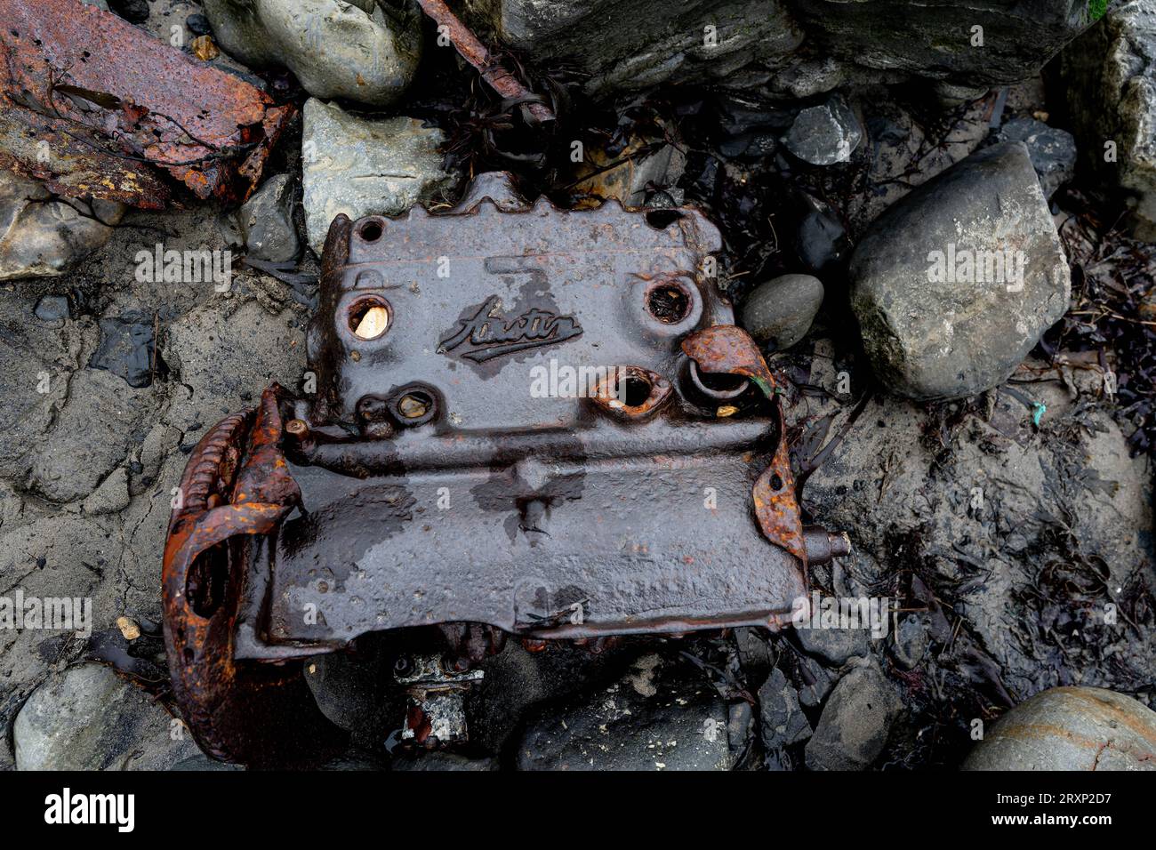 Verrosteter Ford-Motorblock, der an felsigem Ufer zwischen Kieseln und Algen angespült wurde Stockfoto