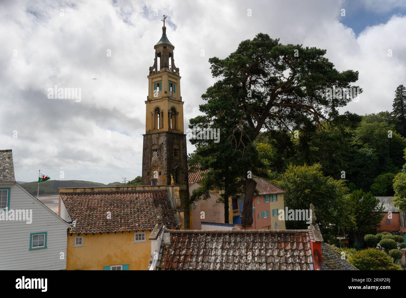 Der historische Glockenturm dominiert das farbenfrohe Dorf Portmeirion mit üppigen Bäumen unter bewölktem Himmel Stockfoto