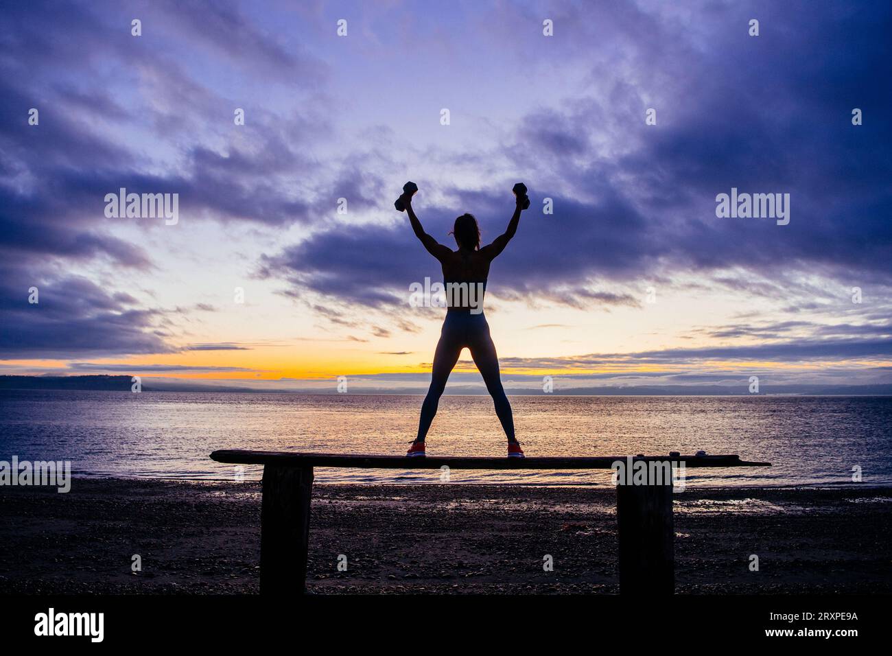 Silhouette einer Frau, die mit Kurzhanteln auf der Küstennbank bei bewölktem Sonnenuntergang trainiert Stockfoto