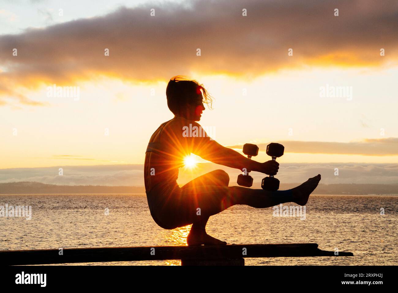 Silhouette einer Frau, die mit Kurzhanteln auf der Küstennbank bei bewölktem Sonnenuntergang trainiert Stockfoto