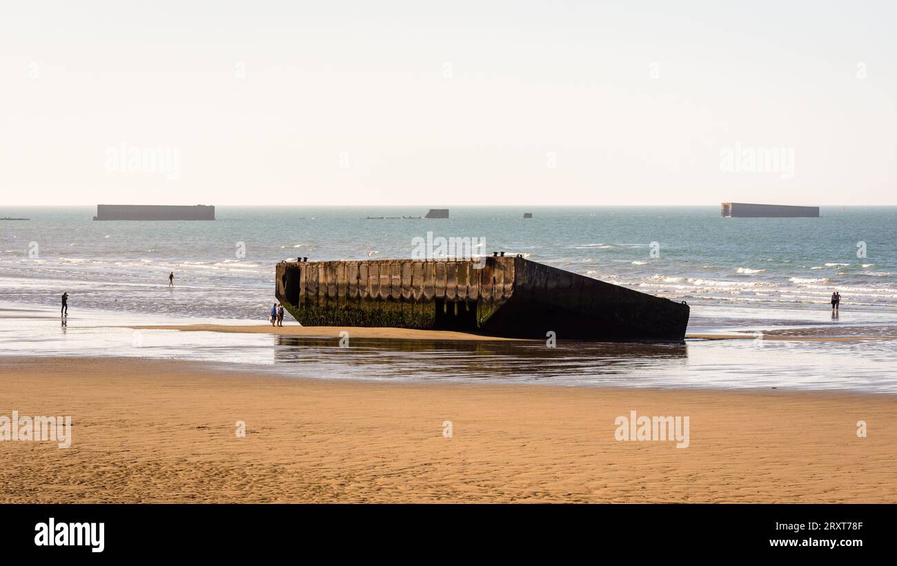 Ein Element des Mulberry-Hafens aus dem Zweiten Weltkrieg, erbaut am Gold Beach in Arromanches, Frankreich, liegt auf dem Sand mit Überresten von Phoenix Caissons in der Ferne. Stockfoto