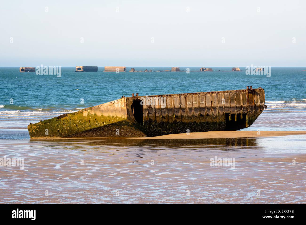 Ein Element des Mulberry-Hafens aus dem Zweiten Weltkrieg, erbaut am Gold Beach in Arromanches, Frankreich, liegt auf dem Sand mit Überresten von Phoenix Caissons in der Ferne. Stockfoto