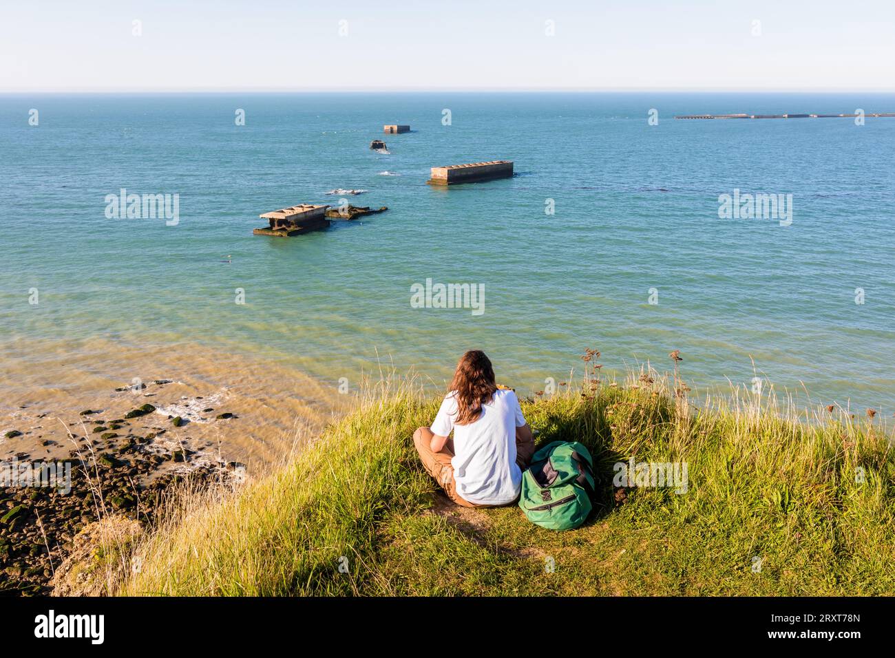 Eine sitzende Frau beobachtet die Überreste des künstlichen Mulberry-Hafens, der nach den Landungen in der Normandie im Zweiten Weltkrieg am Gold Beach in Arromanches, Frankreich, errichtet wurde Stockfoto