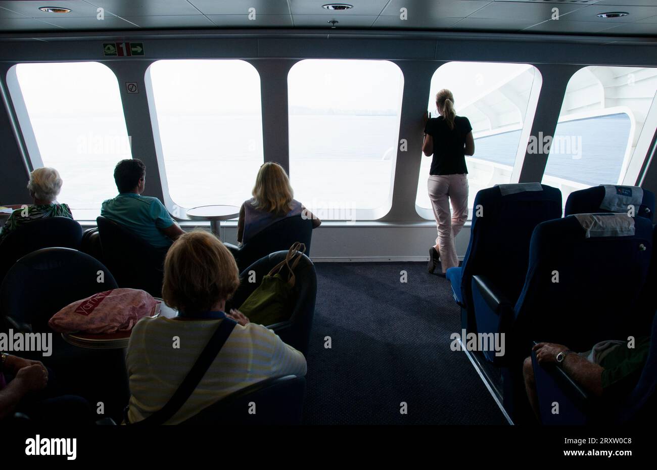 Rückansicht einer Gruppe von Passagieren, die auf dem überdachten Deck einer Fähre sitzen, während sie das Meer durch die großen Fenster der spanischen Küste sehen Stockfoto