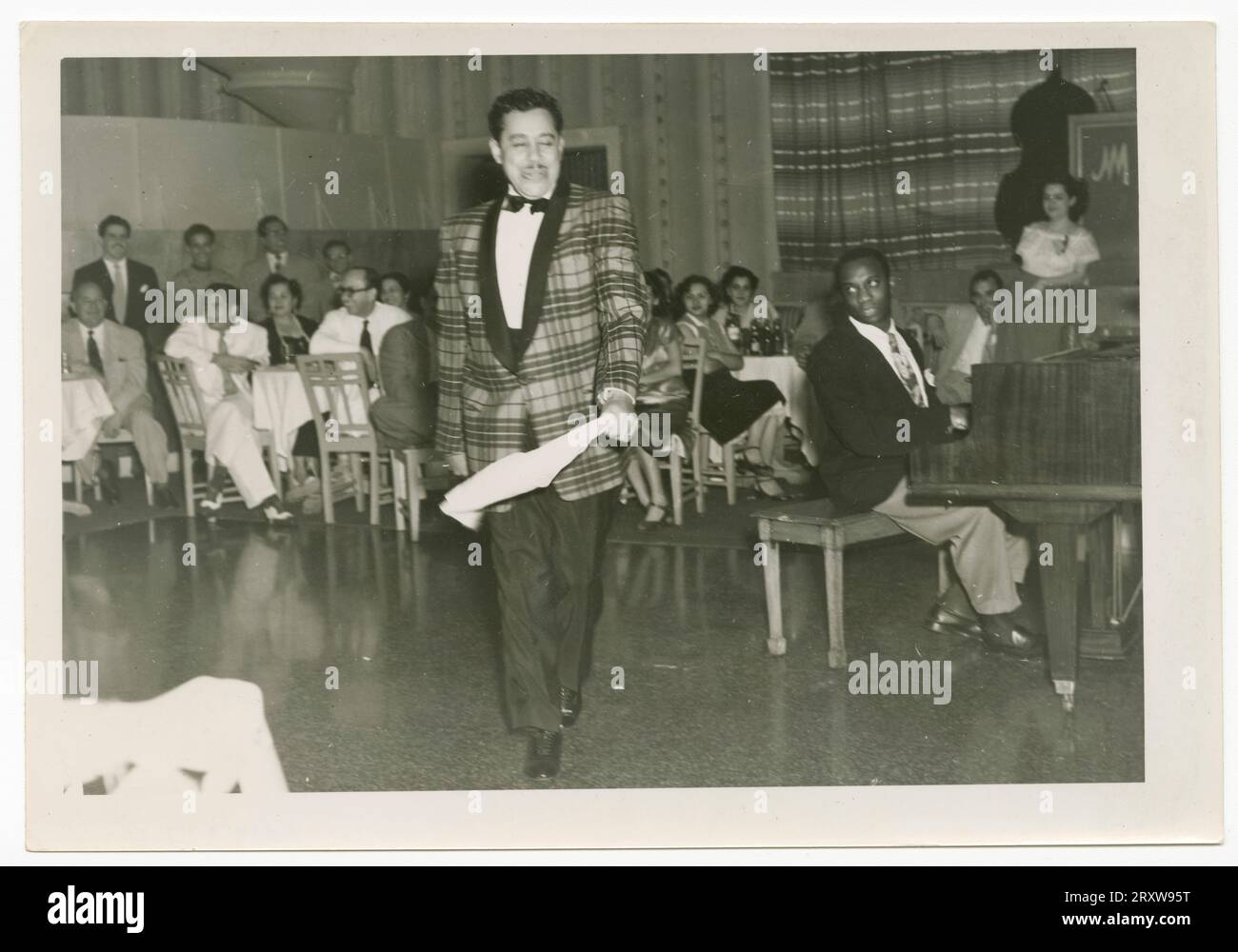S & W-Foto von Cab Calloway, das tanzt, während ein Mann Klavier spielt. Calloway hält ein weißes Taschentuch in seiner PL-Hand und trägt eine karierte Anzugjacke mit dunkler Hose. Eine große Gruppe von Menschen sitzt an Tischen hinter Calloway. Ein gelber Haftnotiz auf der Rückseite des Fotos lautet [Hotel/National/in/Cuba]. Stockfoto