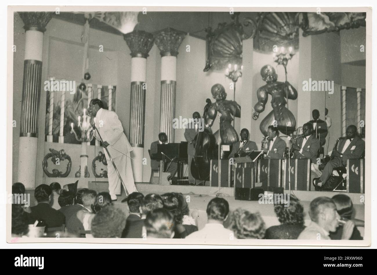 Foto von Cab Calloway auf der Bühne vor seiner Band. Calloway trägt einen hellen Anzug und wird in ein Mikrofon singend dargestellt. Eine große Gruppe sichtbarer Personen, die sitzend auf der Bühne sitzen, entblößt die Bühne. Bandmitglieder auf der Bühne tragen passende dunkle Suiten mit Schleifen. Stockfoto