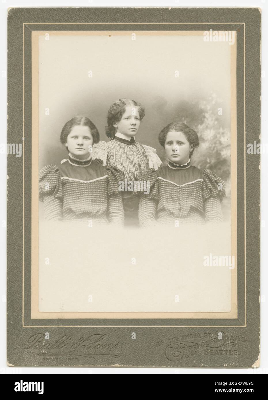 Ein Schwarzweißfoto von drei Mädchen, die im Ball & Sons Studio in Seattle, WA, fotografiert wurden. Das Foto befindet sich auf einer dunklen Schrankkarte, die mit dem Ball & Sons-Logo bedruckt ist, und befindet sich an der Unterseite der Vorderseite. Das Trio wird von der Hüfte nach oben fotografiert, zwei Mädchen sitzen und das dritte steht zwischen und hinter den anderen beiden. Die beiden sitzenden Mädchen tragen identische dunkelfarbige Kleider und das dritte Mädchen trägt ein helleres Kleid mit Spitzendekorationen auf dem Ärmel. Die Mädchen haben alle dunkle Haare, die zurückgezogen wurden. Es gibt eine Inschrift in Bleistift o Stockfoto
