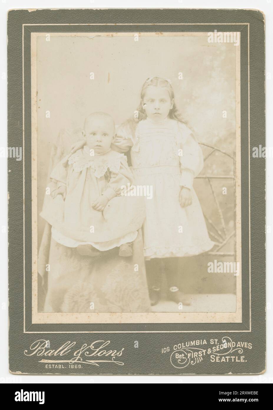 Ein Schwarzweißfoto von zwei kleinen Kindern, das im Ball & Sons Studio in Seattle, WA, fotografiert wurde. Das Foto befindet sich auf einer dunklen Schrankkarte, die mit dem Ball & Sons-Logo bedruckt ist, und befindet sich an der Unterseite der Vorderseite. Auf dem Foto sitzt ein Kleinkind in einem drapierten Stuhl, und ein älteres Kind steht links neben dem Kleinkind mit der Hand um die Schulter des Babys. Beide Kinder hatten helle Kleider. Stockfoto