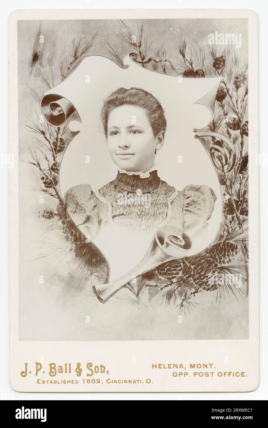 Eine Schwarzweißfotografie einer Frau, die im J. P. Ball & Son Studio in Helena, MT, fotografiert wurde. Das Foto ist auf einer hellen Schrankkarte montiert, auf der der Name und die Position des Fotostudios auf der Vorderseite entlang der Unterseite aufgedruckt sind. Das Porträt ist von einem dekorativen Rand aus gewelltem Scrollpapier und Kiefernzweigen umgeben. Das Mädchen trägt ein gemustertes Kleid mit Rüschenkragen und Medaillon. Sie hat dunkles Haar, das zurückgesteckt ist. Auf der Rückseite der Karte befindet sich eine Bleistiftbeschriftung. Stockfoto