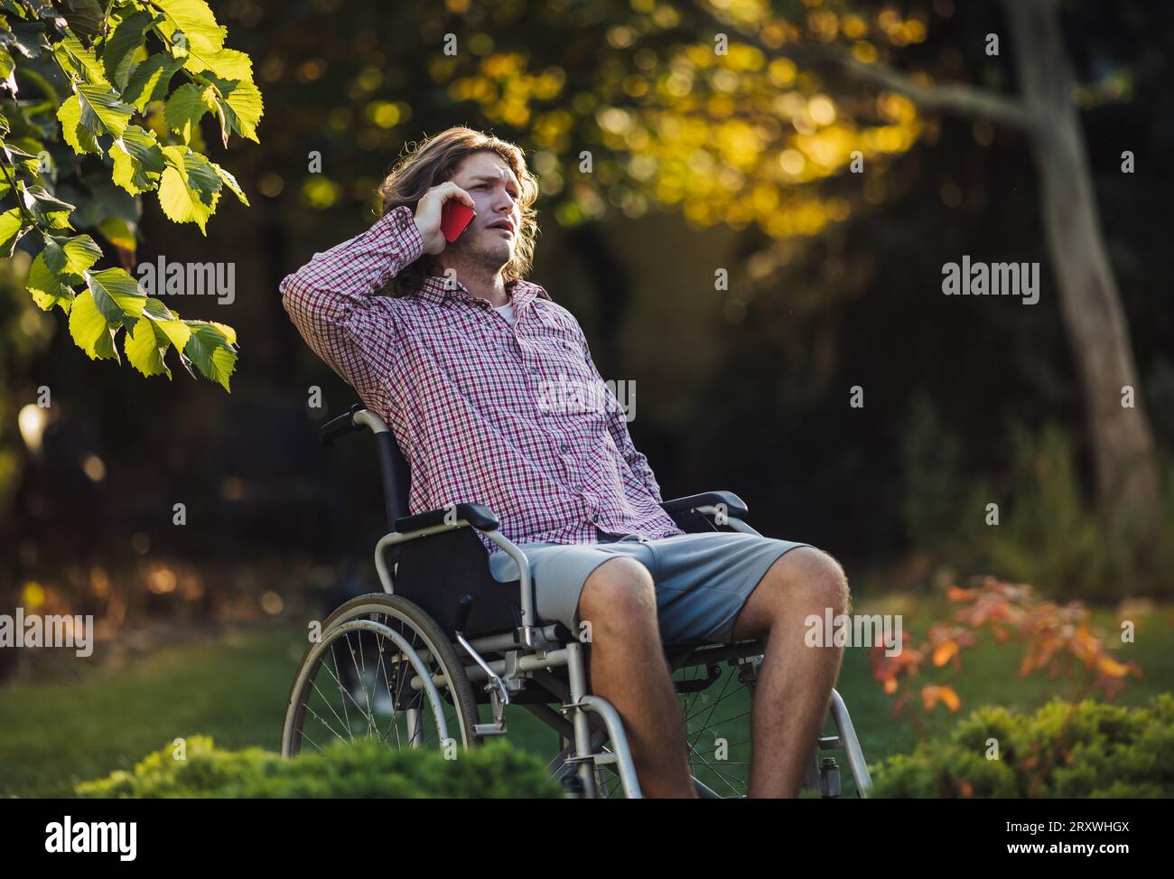 Unglücklicher junger Mann im Rollstuhl, der auf dem Handy im Park spricht Stockfoto