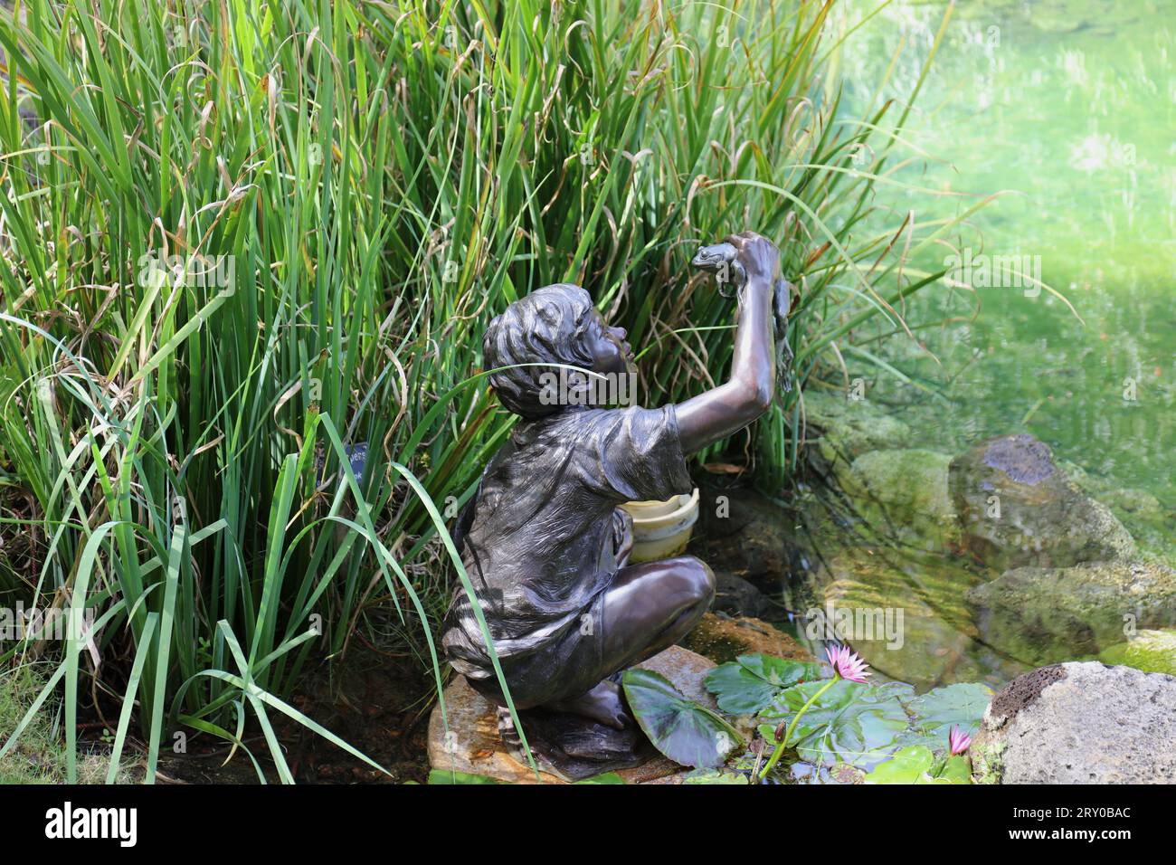 Eine Bronzeskulptur eines kleinen Jungen, der einen Frosch am Ende eines Teichs zwischen Wasserpflanzen in den Na Aina Kai Botanical Gardens in Kauai, Hawaii, USA hält Stockfoto