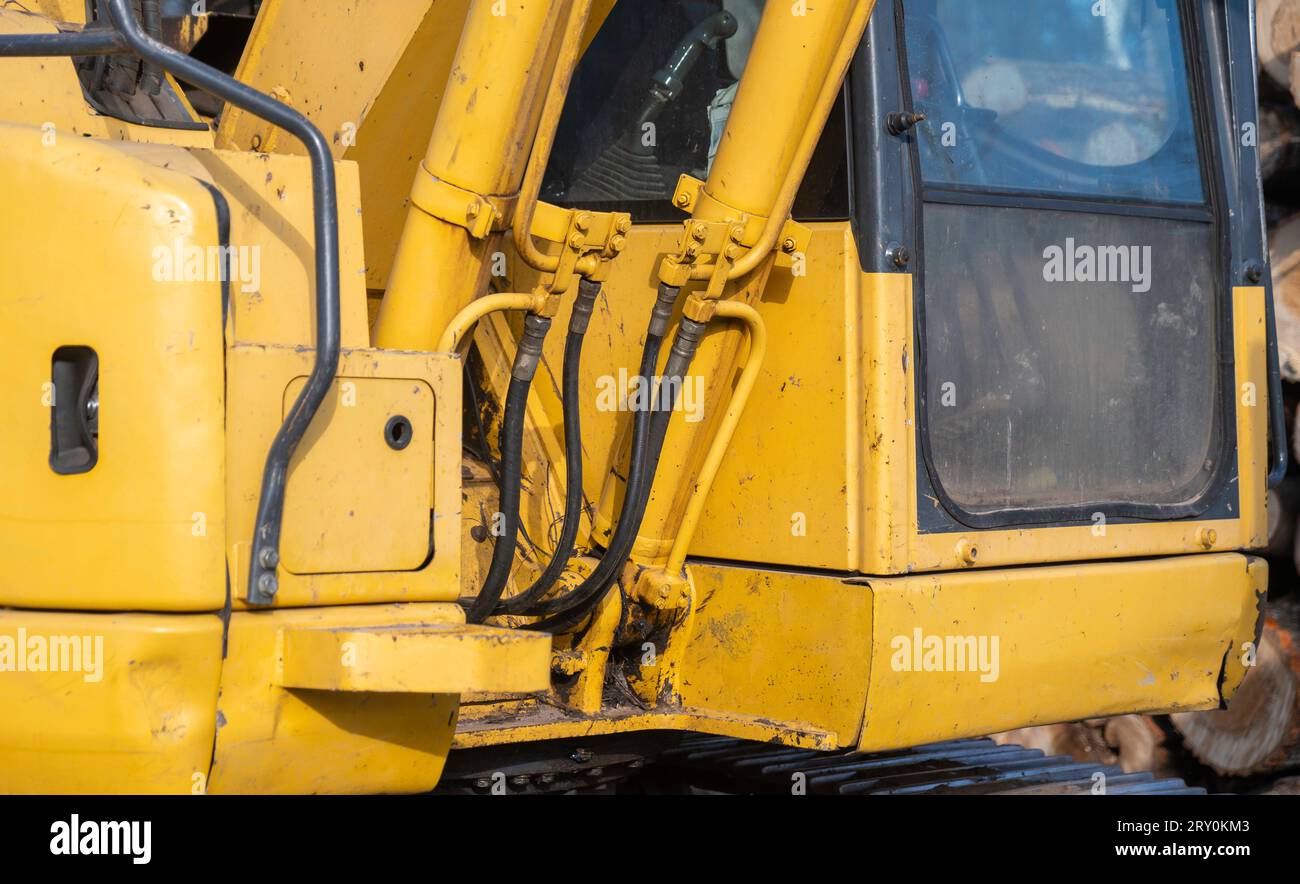 Nahaufnahme eines funktionierenden, gebrauchten, schmutzigen gelben Baggers, zentriert auf Hydraulikschläuchen und -Leitungen neben dem Fenster der Fahrerkabine. Stockfoto