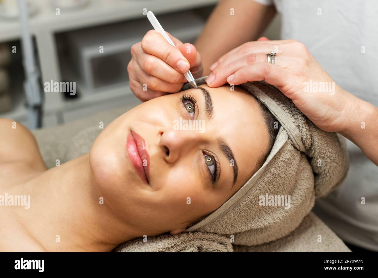 Große inhaltliche Dame mit Handtuch auf dem Kopf, die auf dem Bett liegt, während anonyme Kosmetikerin im Schönheitssalon Augenbrauen zupft Stockfoto