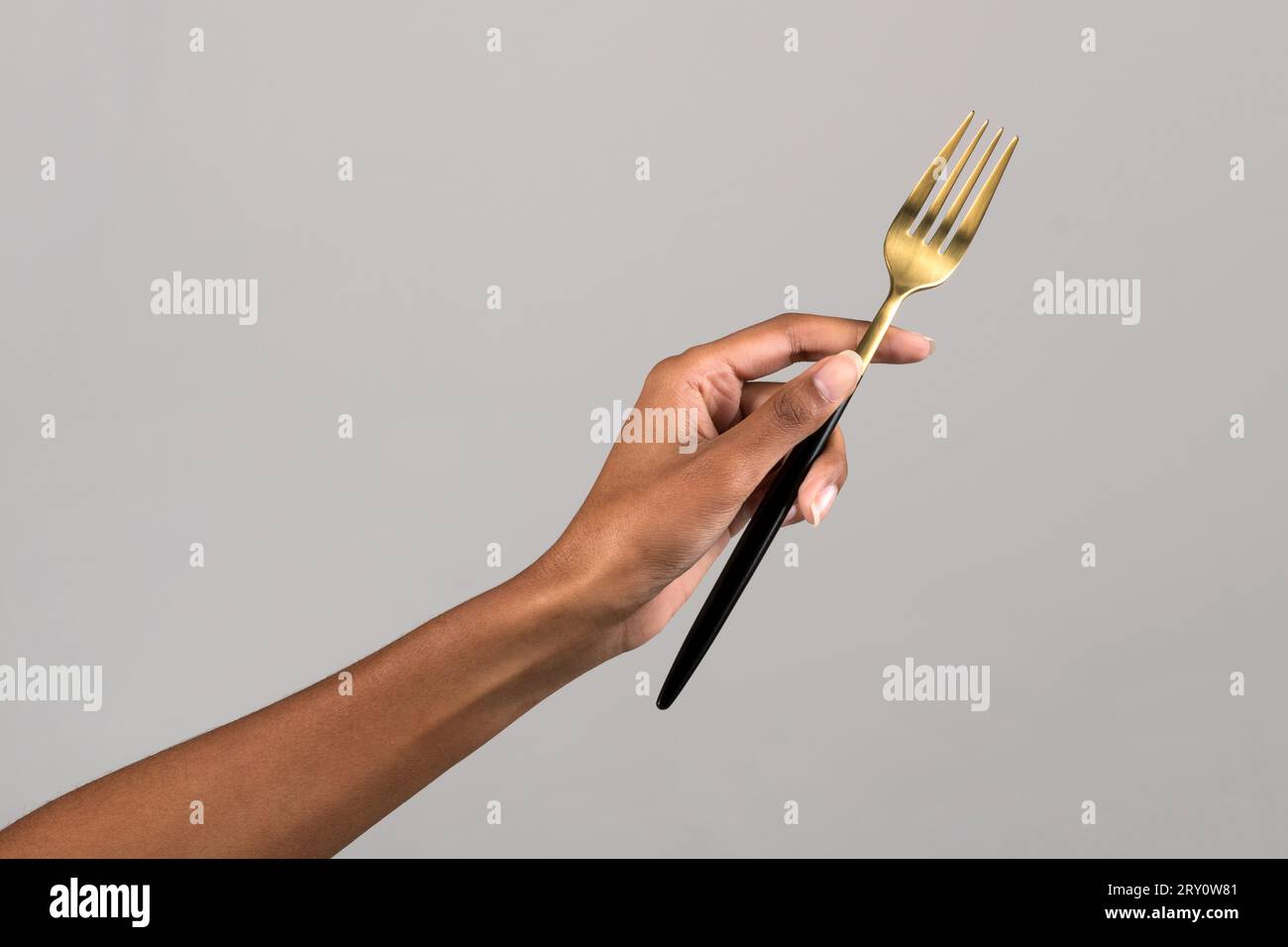 Die anonyme Hand einer afroamerikanischen Frau mit goldfarbener Gabel vor grauem Hintergrund Stockfoto