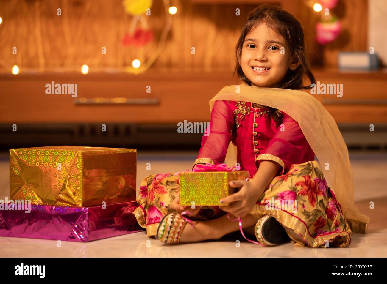 Glückliches kleines indisches Mädchen, das Geschenkboxen in der Hand hält, um Diwali zu feiern, während es auf dem Boden sitzt, Raksha Bandhan, Bhai Dooj Concept. Stockfoto