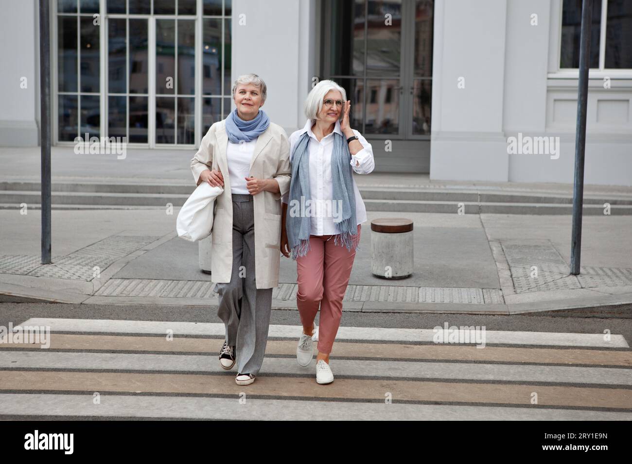 Zwei ältere Frauen mit grauen Haaren in modischer, stylischer Kleidung, die auf der Straße spazieren gehen und die Straße überqueren. Seniorinnen im Freien. Stockfoto
