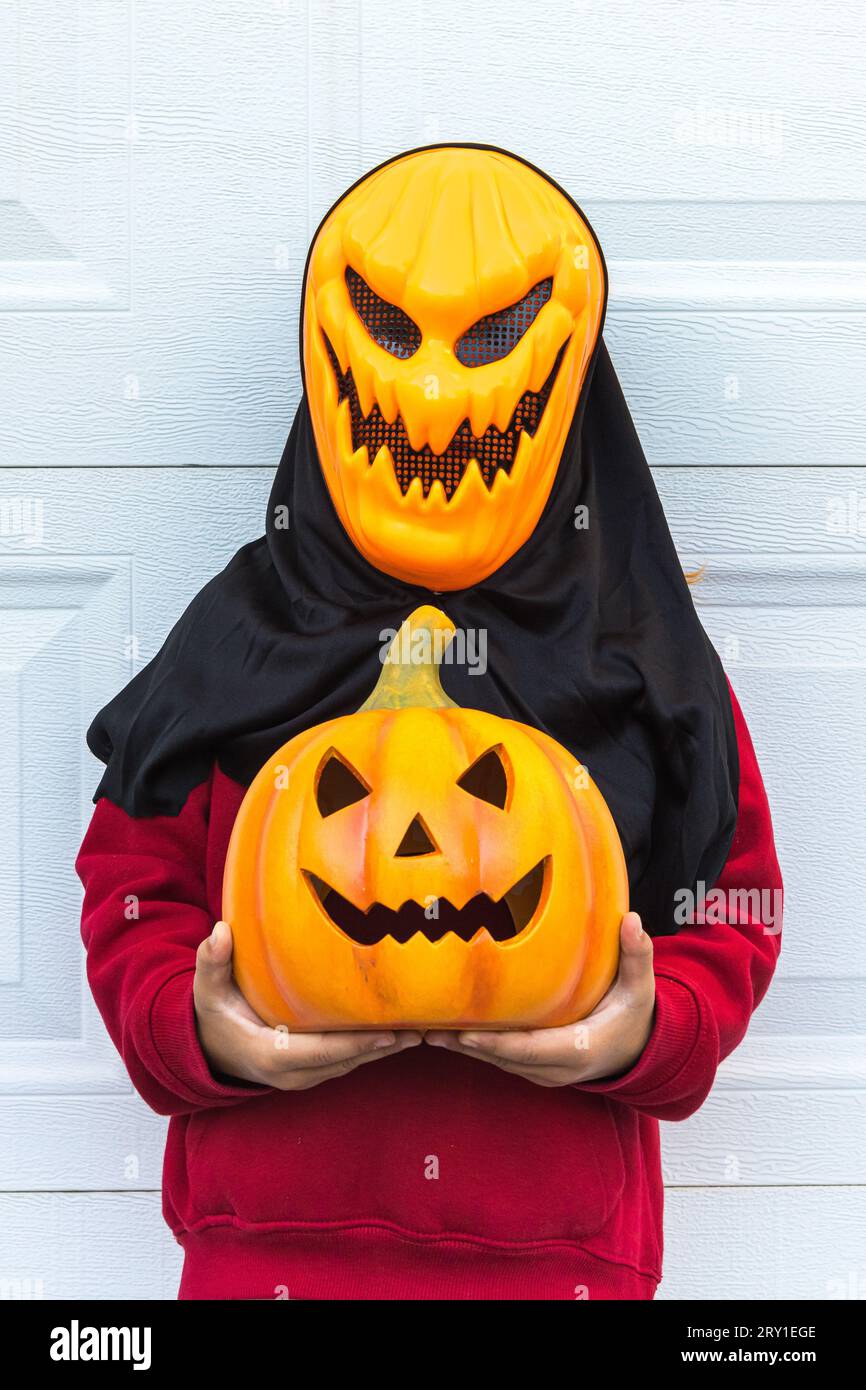 Ein Kindermädchen, das eine gruselige Halloween-Kürbismaske trägt, einen Kürbis hält, mit der Hand erschreckt, über einem weißen Garagentor. Das Konzept des Feierns, Cou Stockfoto