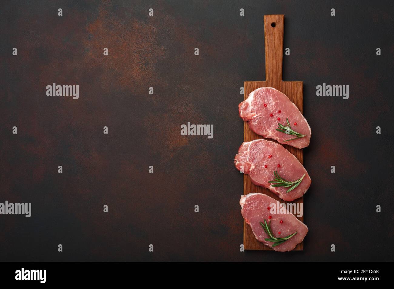 Rohes Schweinesteak auf Schneidebrett mit Rosmarin und roter Paprika auf rostrotem braunem Hintergrund. Draufsicht mit Platz für Ihren Text. Stockfoto