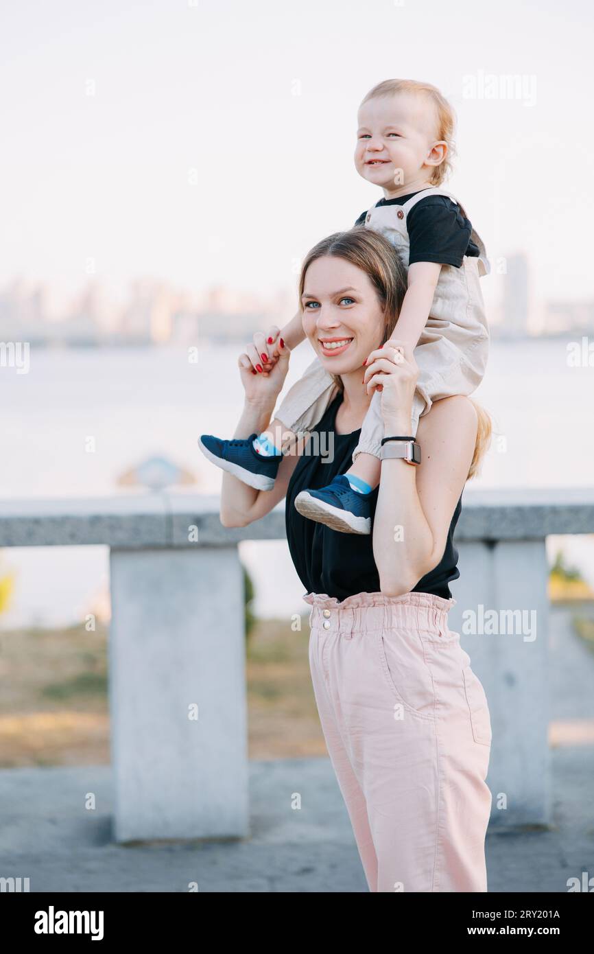 Glückliche junge Mutter, die ihren kleinen Jungen auf den Schultern trägt, während sie in der Stadt vor dem Hintergrund des Flusses und der Stadtlandschaft spaziert. Stockfoto