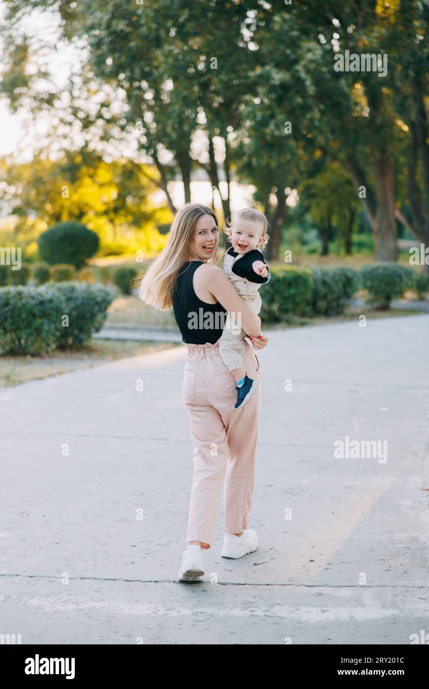 Glückliche junge Mutter hat Spaß, läuft und schaut mit ihrem Kleinkind zurück, während sie im Park spaziert. Stockfoto