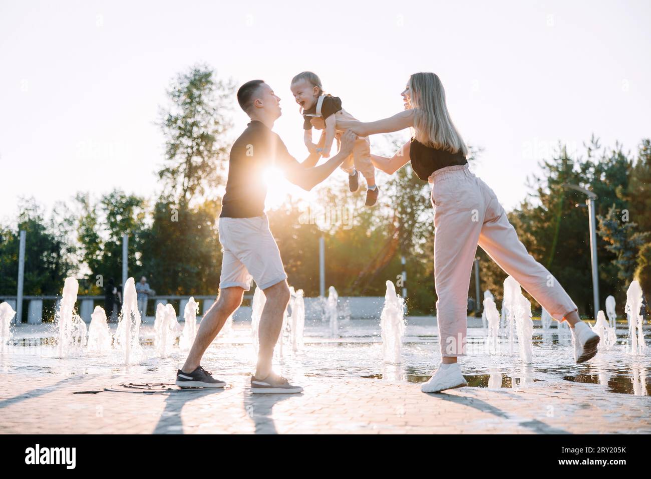 Glückliche Eltern, die mit seinem Kleinkind spielen, während sie im Park vor dem Hintergrund des Brunnens spazieren. Hintergrundbeleuchtung. Stockfoto