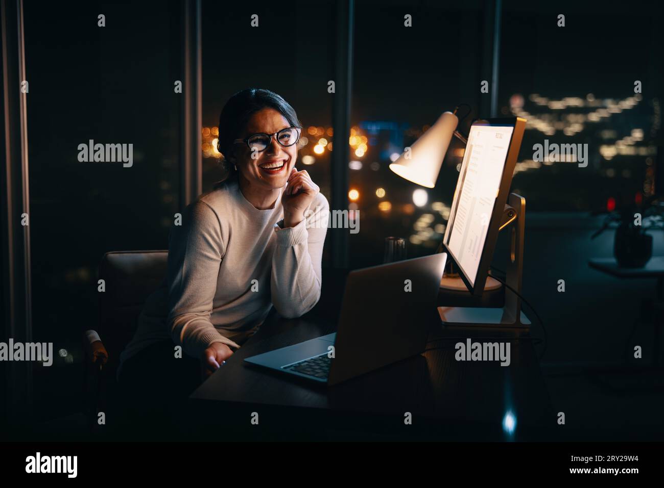 Glückliche Geschäftsfrau lächelt vor der Kamera, während sie lange in ihrem Büro arbeitet. Professionelle Frau, die lange Stunden einsetzt, um ihre Produktivität zu steigern und Stockfoto