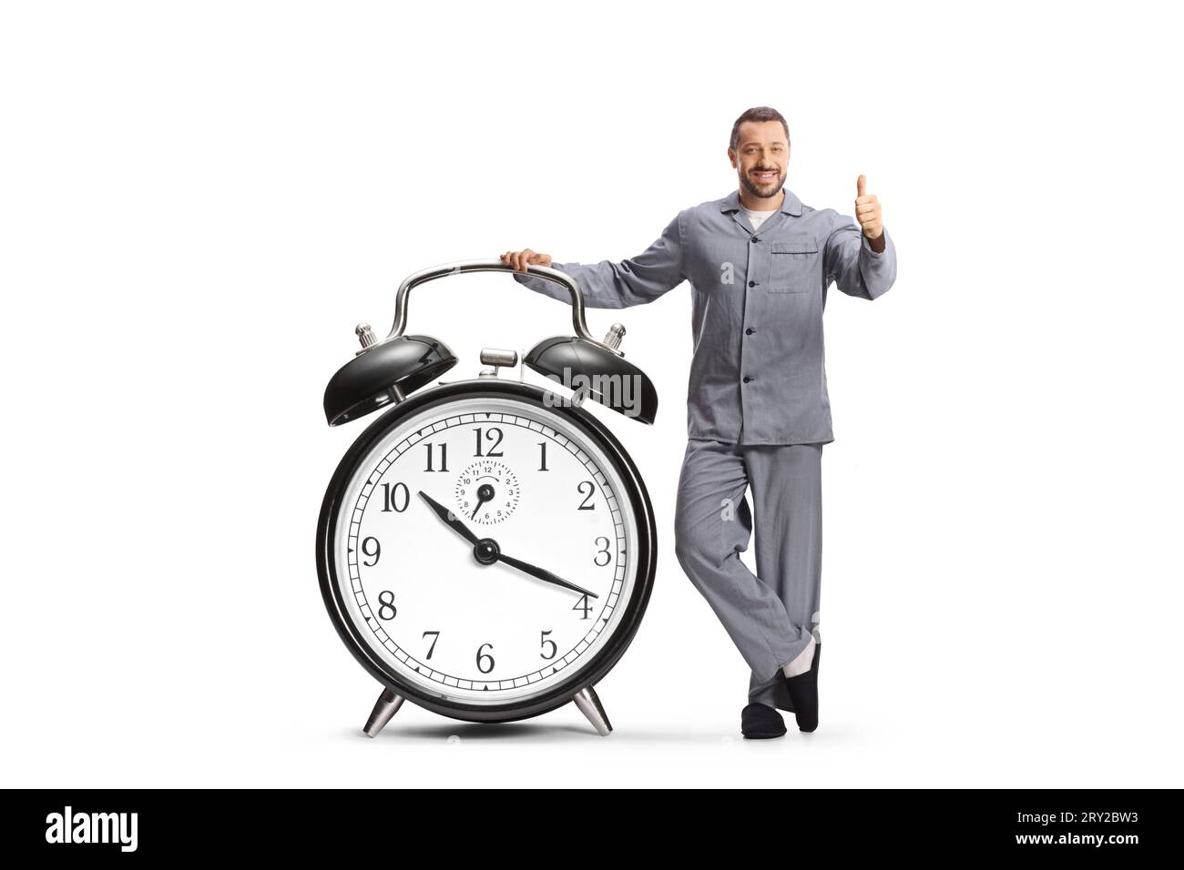 Mann im Pyjama, der sich auf einen großen Wecker lehnt und die Daumen auf weißem Hintergrund bewegt Stockfoto
