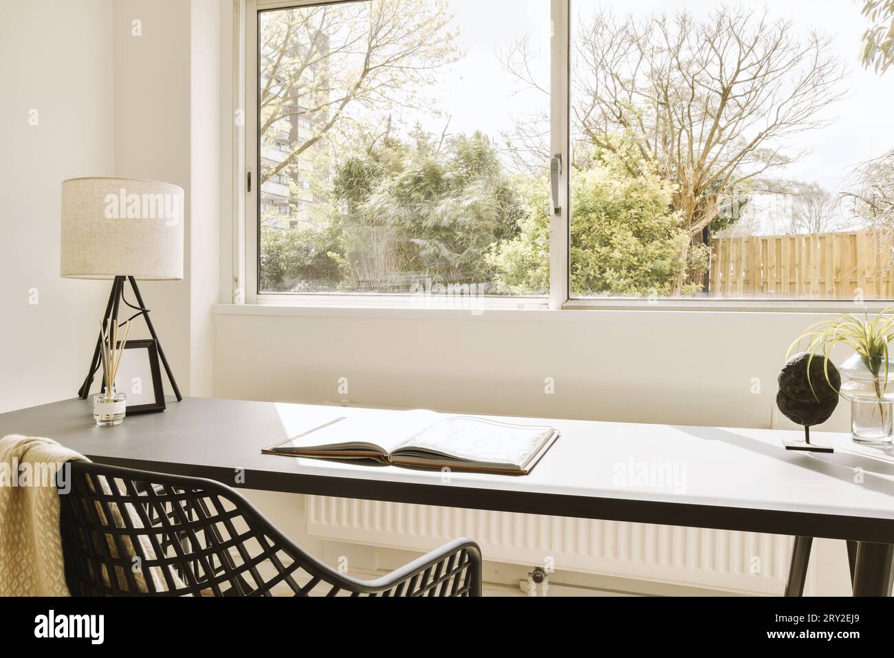 Bücher mit Lampe und kleinen Pflanzen auf dem Schreibtisch vor einem großen hellen Fenster im Home Office in einem modernen Apartment Stockfoto