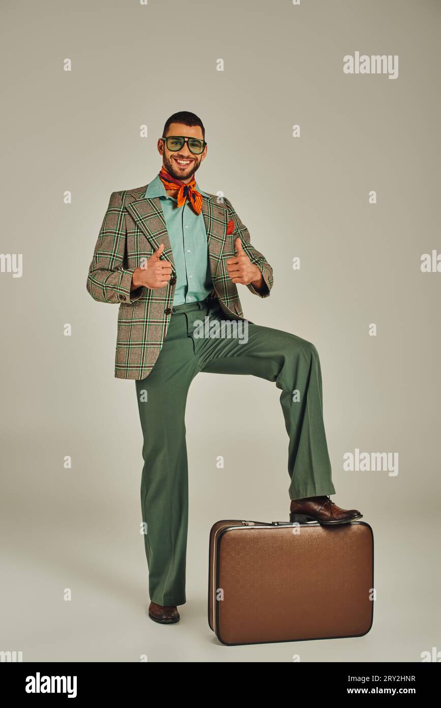 Glücklicher Mann in Retro-Kleidung und Sonnenbrille mit Daumen nach oben in der Nähe von Vintage-Koffer auf Grau Stockfoto