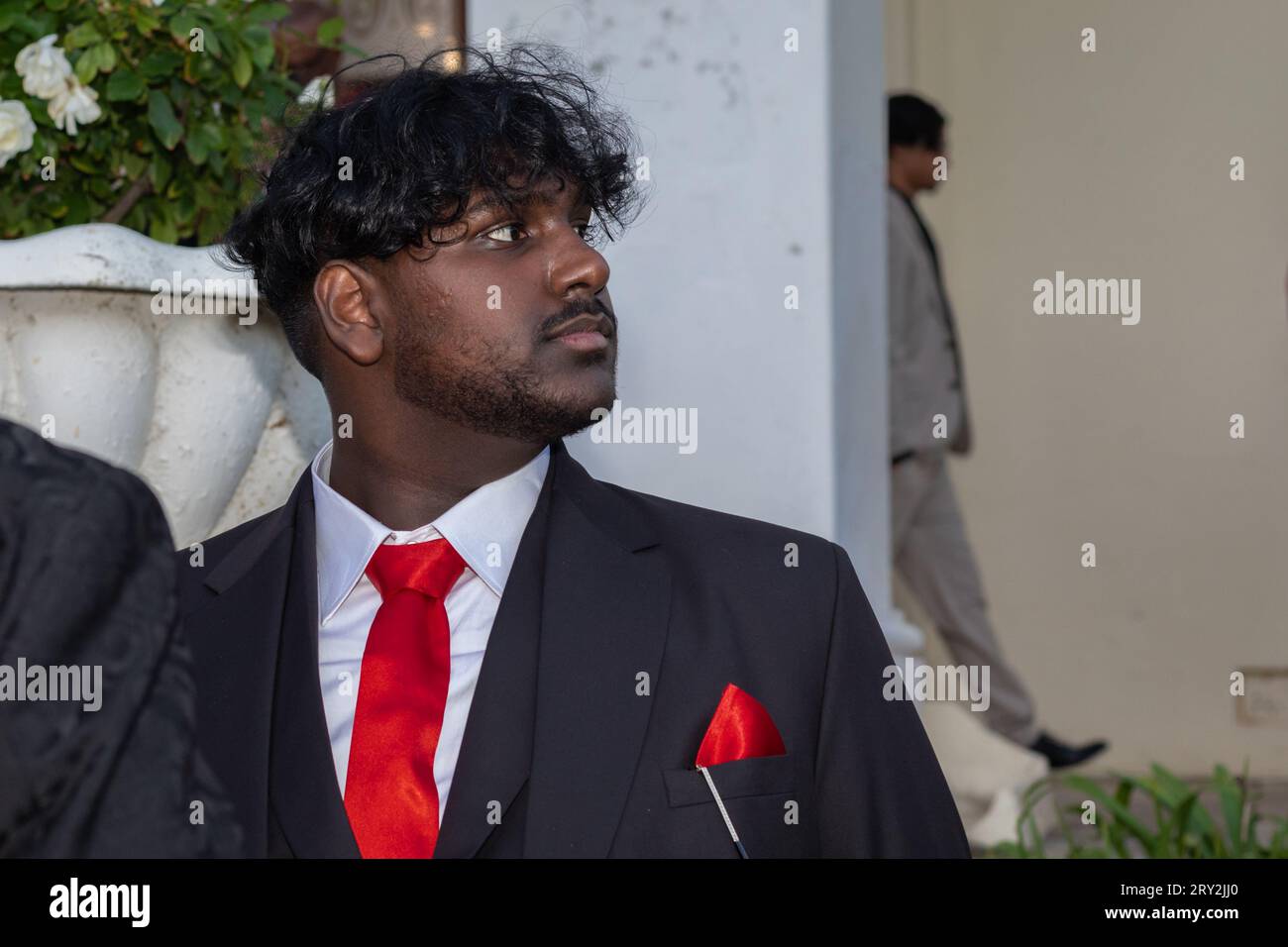 In einer Nahaufnahme blickt ein junger indischer Mann mit roter Krawatte und rotem Anzug zur Seite, während ein anderer Mann hinter einer Säule im Hintergrund heraustritt Stockfoto