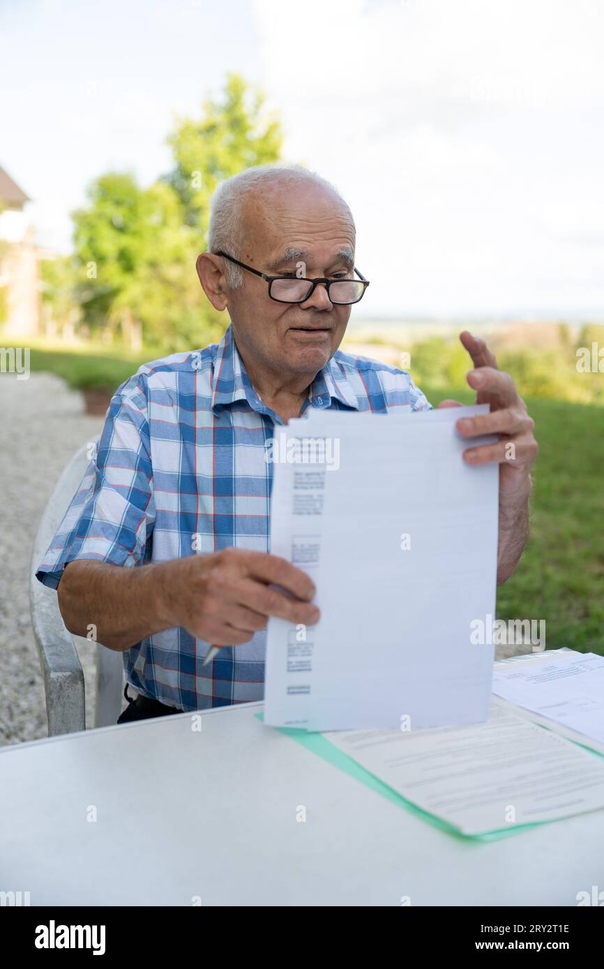 Ein älterer Mann sitzt draußen an einem Schreibtisch, arbeitet mit Dokumenten, füllt seine medizinischen Formulare aus, trägt eine Brille, hält einen Stift. Stockfoto