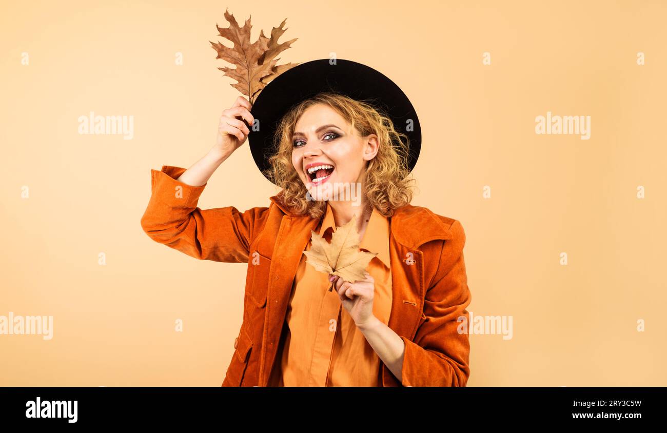 Modefrau mit schwarzem Hut und Herbstblättern. Lächelndes weibliches Modell mit perfektem Make-up in modischer Herbstmode. Stilvolles Mädchen im orangefarbenen Mantel mit Stockfoto