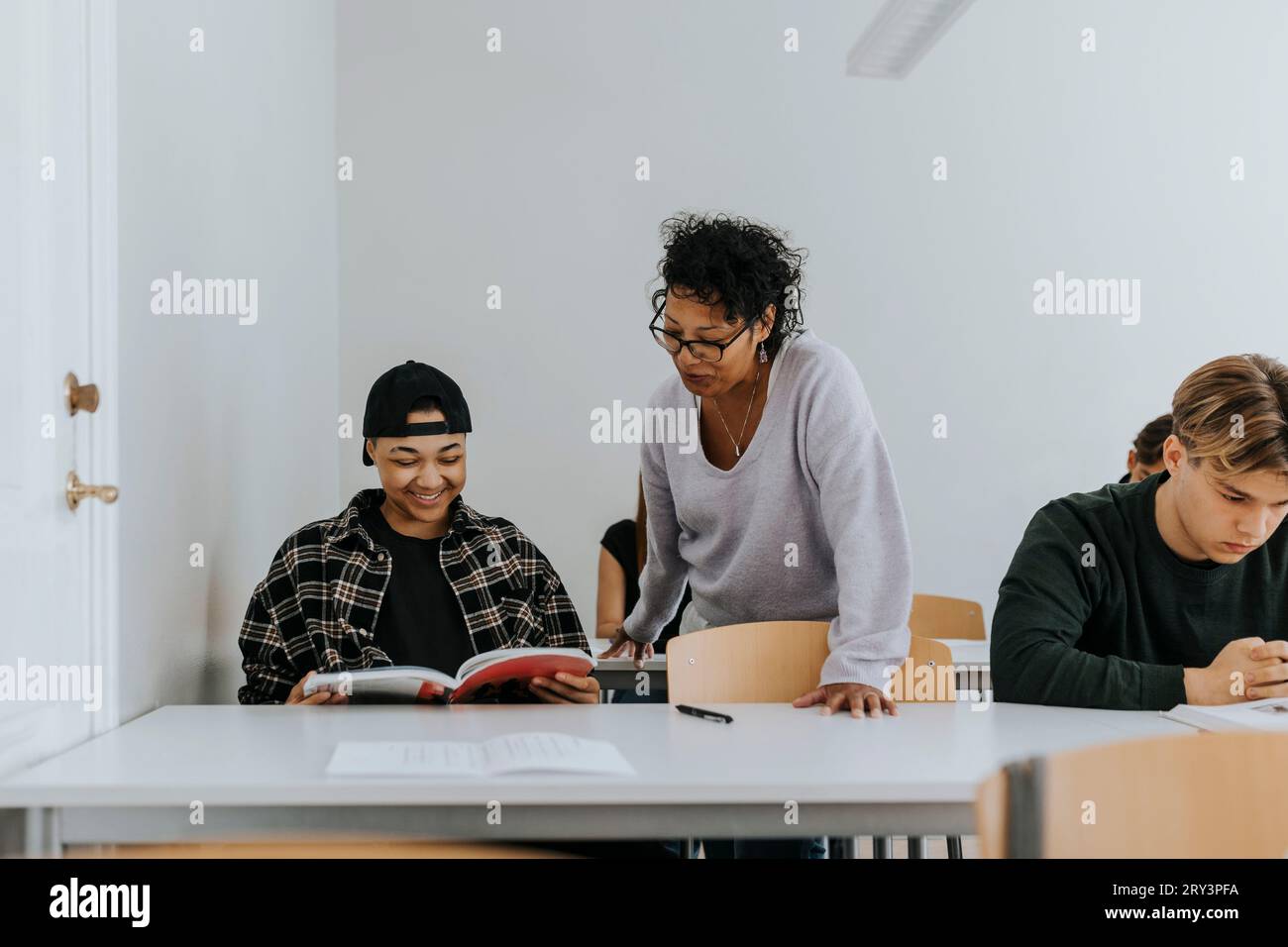 Der Lehrer steht neben einem lächelnden männlichen Schüler, der im Klassenzimmer am Schreibtisch sitzt Stockfoto
