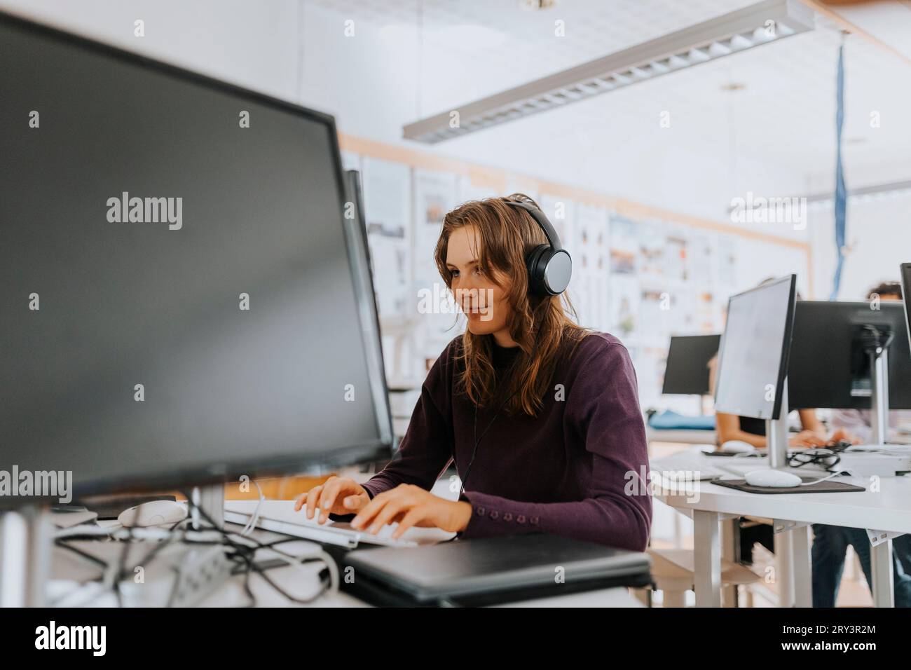 Junge Frau, die durch Kopfhörer hört, während sie im Schullabor am Computer arbeitet Stockfoto