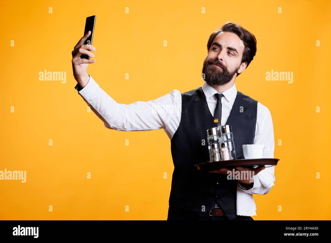 Junger Kellner, der Fotos im Studio macht und mit dem Handy ap dumme Bilder von ihm mit der Platte in der Hand macht. Butler, der im Restaurant arbeitet, nimmt Selfies vor gelbem Hintergrund. Stockfoto
