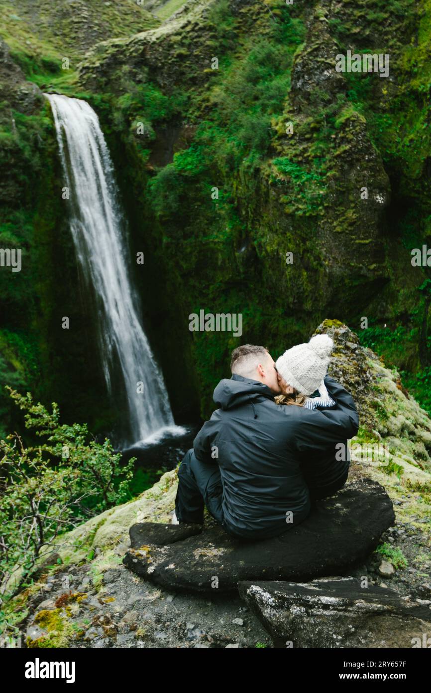 Verheiratete Paare küssen mit Blick auf den Wasserfall, den grünen, üppigen Klippen hinunter Stockfoto